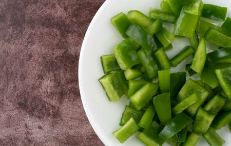 Top view of chopped green bell peppers on a white plate atop a maroon counter top.