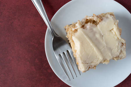 Top view of a piece of homemade banana bread with vanilla icing on a plate with a fork atop a maroon...