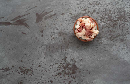 Top view of pancetta sliced into cubes in a small bowl offset on a gray table illuminated with...