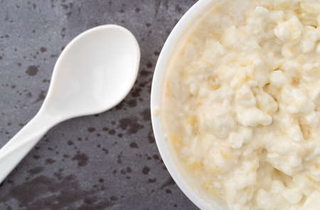 Overhead close view of a container of pineapple cottage cheese with a spoon to the side on a gray table illuminated with natural lighting.の写真素材