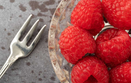 Overhead close view of freshly picked raspberries in a small glass bowl with a fruit fork to the side on a gray background illuminated with natural lighting.の写真素材