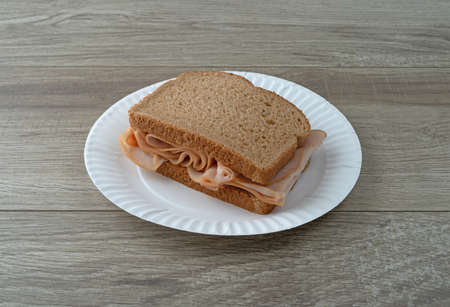 Sandwich with thin sliced smoked turkey on wheat bread on a white plate atop a table illuminated with natural light.の写真素材