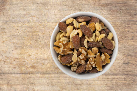Assorted nuts in a white bowl atop an old wood cutting board top view.