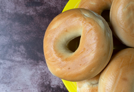 Top view of a group plain bagels on a large yellow plate atop a red mottled tabletop illuminated with natural lighting.の写真素材