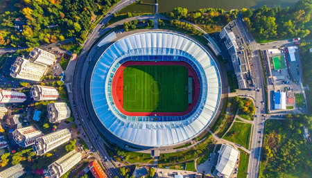 An aerial drone shot captures a large, modern stadium with a vibrant green field amidst a dense urban landscape of tall residential buildings, roads. AI-generated contentの素材