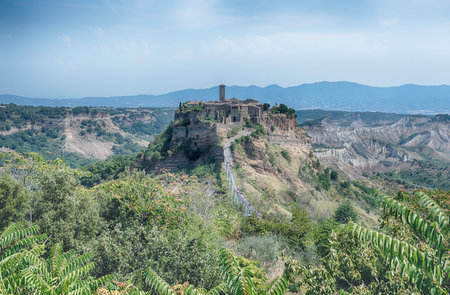A historic village is perched atop a steep, eroded hill, connected by a long pedestrian bridge. Lush green vegetation and barren badlands surround the iconic Italian landmark under a clear sky.の写真素材