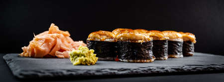 Panoramic photo of served warm sushi rolled in nori sheets with ginger and wasabi on a black stone...