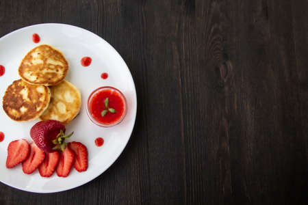 Three pancakes with cheese and strawberries and sauce on a white dish against wood background. Delicious cheese pancake and strawberries for the breakfast or lunch. Tasty meal with berries.の写真素材