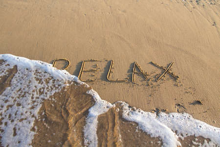 Word RELAX wrote on the sand against background. Message says "relax" in the sand on the beach with sea foamの写真素材