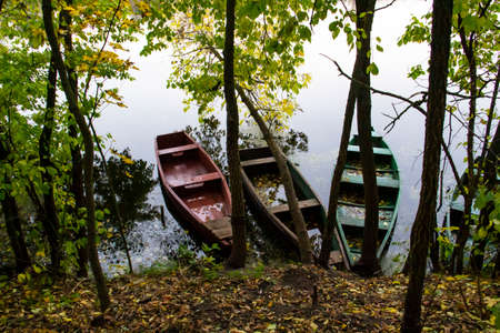 Empty boats are tied up by the shore in front of the river. Autumn landscape with trees and boatsの写真素材