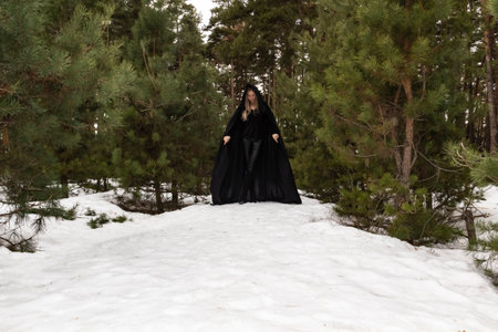 Blonde woman in a black cloak with a hood in winter in a pine forestの写真素材