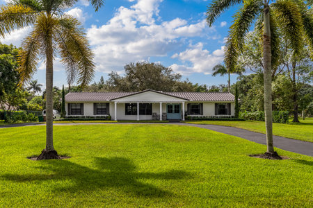 Beautiful facade of a white house with black tiles, with a park with short grass, trees and palm trees, blue sky, drivewayの写真素材