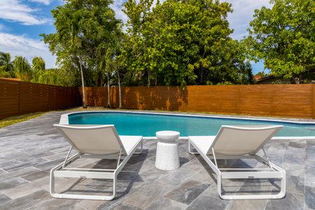 point of view of turquoise pool area with two sun loungers, wooden fence, palms and trees, blue skyの写真素材