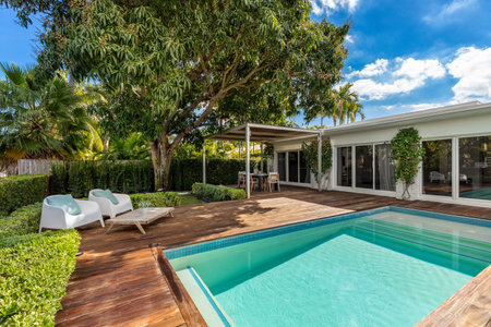 Beautiful patio with dark wood floor, swimming pool, bushes, trees, wooden coffee table lounge chairs, blue sky with clouds in Biscayne Pointの写真素材