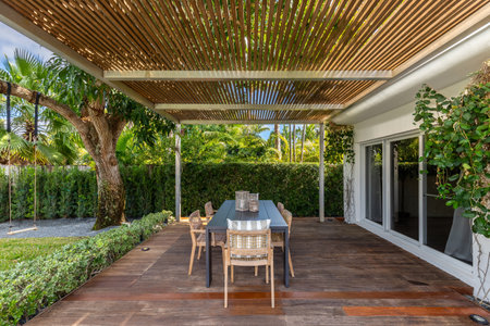 Beautiful patio with dark wood floor, swimming pool, bushes, trees, wooden coffee table lounge chairs, blue sky with clouds in Biscayne Pointの写真素材
