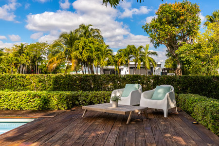 Beautiful patio with dark wood floor, swimming pool, bushes, trees, wooden coffee table lounge chairs, blue sky with clouds in Biscayne Pointの写真素材