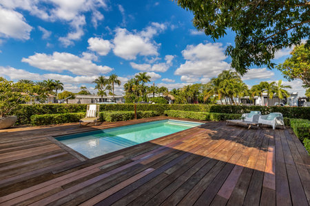 Beautiful patio with dark wood floor, swimming pool, bushes, trees, wooden coffee table lounge chairs, blue sky with clouds in Biscayne Pointの写真素材