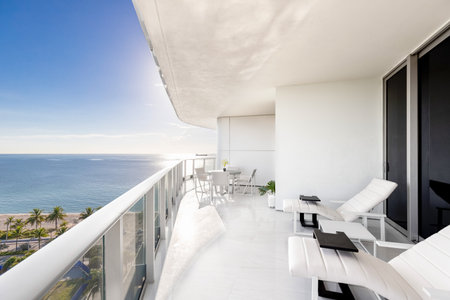 Shot of white balcony with view of fort lauderdale beach in Central Beach neighborhood, outdoor furniture, chairs and table, palms and tropical vegetation, turquoise sea and blue sの写真素材