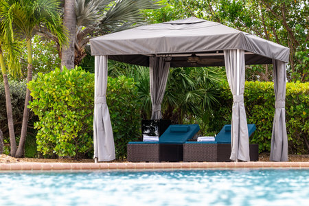 Point of view from lounge chairs with rolled towel in gazebo with fan, swimming pool, surrounded by greenery and tropical plants, summer weather, blue skyの写真素材