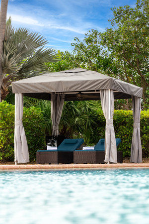 Point of view from lounge chairs with rolled towel in gazebo with fan, swimming pool, surrounded by greenery and tropical plants, summer weather, blue skyの写真素材