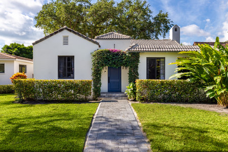Front of a colonial style and modern white house, in the Miami shore of Miami-Dade, with a privet arch at the door, driveway, sidewalk, short grass, tiled roof, trees and palms, whiteの写真素材
