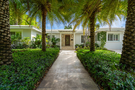 facade of a modern and luxurious colonial-style house in the city of Coral Gables, Miami-Dade, USA, with large tropical vegetation around it, open garage, main entrance, sidewalk,の写真素材