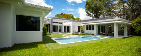 Backyard of elegant and modern house, located in Glenvar Heights, in Miami-Dade, white walls, black tiles, glass doors, short cepes, T-shaped pool, trees and palm trees around, sunの写真素材