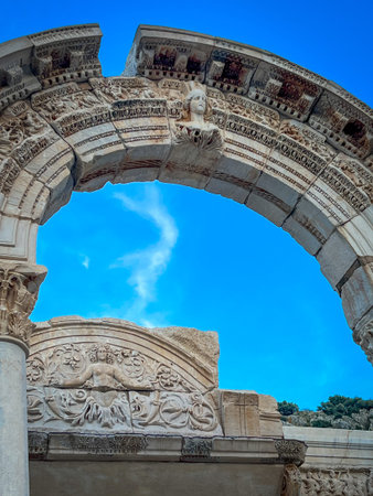 Towering marble archway from Ephesus' Temple of Hadrian showcases weathered reliefs including a protruding female bust and reclining figures against fractured stone. Clear blue sky with faint clouds pierces through the central opening, emphasizing intricate Roman detailing and classical proportions.の写真素材