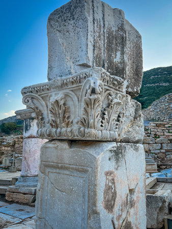 Low-angle perspective emphasizes the intricate acanthus leaves and sphinx figures chiseled into the fragmented column's capital, set against a vibrant blue sky. Cracked stone textures and surrounding debris on tiled pavement evoke enduring Roman grandeur with terraced hillside backdrop.の写真素材