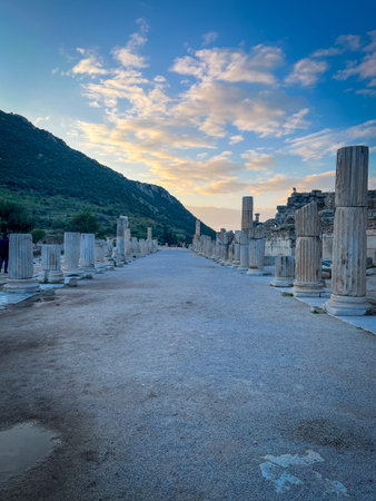 Fluted marble columns rise symmetrically along a straight stone path, their weathered surfaces catching golden evening rays amid scattered clouds. Crumbling ruins perch on one side while lush green mountains loom in the distance, shadows elongating across reflective wet pavement.の写真素材