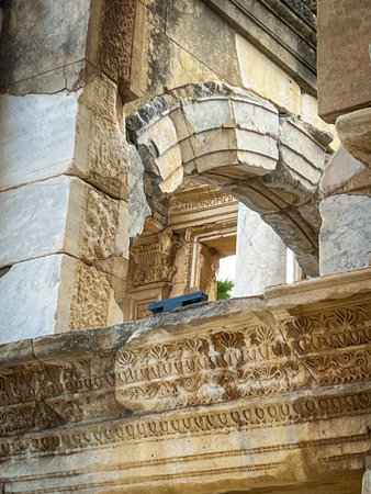 Curved broken pediment arches prominently above columns etched with intricate acanthus scrolls and motifs on the weathered stone facade of the Library of Celsus. Subtle green foliage clings to the ledge beside a blue sign, accentuating the aged textures and historical depth.の写真素材