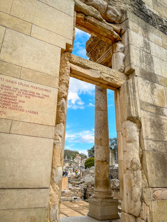 Latin inscriptions honor Tiberius Julius Celsus Polemaeanus atop the weathered stone entablature, with Corinthian columns rising on pedestals through the grand Roman doorway. Blue sky and distant ruins with tiny figures peek beyond, capturing the textured beige facade in bright daylight.の写真素材