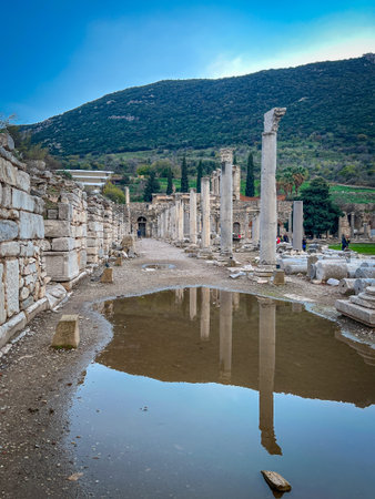 Shallow puddles on the dirt path reflect the weathered stone columns of Ephesus' Roman Agora,...