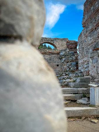 Weathered white stone block dominates the foreground, framing a crumbling red-brick archway with visible green trees beyond and ascending stone steps scattered with rubble. Blue sky dotted with fluffy clouds adds depth to the ancient masonry remnants.の写真素材