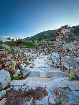 Uneven slabs of weathered marble form a pathway flanked by crumbling stone walls and scattered column bases under a vivid blue sky. Modern yellow lanterns punctuate the scene amid green moss and distant rocky outcrops topped with ancient debris.の写真素材