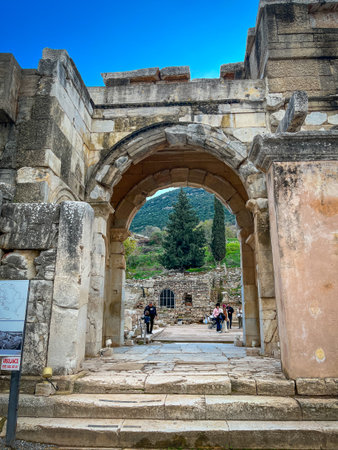 Weathered limestone archway of Mazeus and Mithridates rises boldly in ancient Ephesus, channeling views of cypress trees, rugged hills, and scattered ruins through its grand opening under vivid blue skies. Tourists climb broad stone steps along the historic path blending Roman grandeur with natural vistas.の写真素材