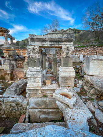 Fragmented stone gateway with weathered fluted columns and collapsed pediment rises amid tumbled blocks and drums on a rocky hillside terrace. Bare-branched trees frame the bright blue sky with wispy clouds, casting soft shadows on textured beige and white stone surfaces.の写真素材
