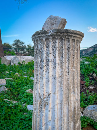 Jagged white stone block teeters precariously on the weathered flutes of a tall marble column rising from green-shrouded rubble. Scattered ancient debris and rocky hillside recede into a vivid blue sky, capturing enduring classical decay.の写真素材