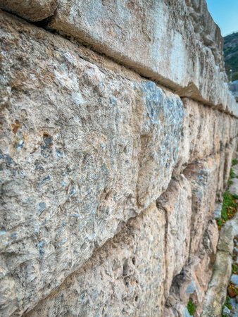 Tight close-up reveals irregularly shaped limestone blocks stacked in precise horizontal courses, showcasing cracks, erosion, and subtle moss at the base. Hazy sky backdrop enhances the textured surface and earthy beige-gray patina of this ancient retaining wall.の写真素材