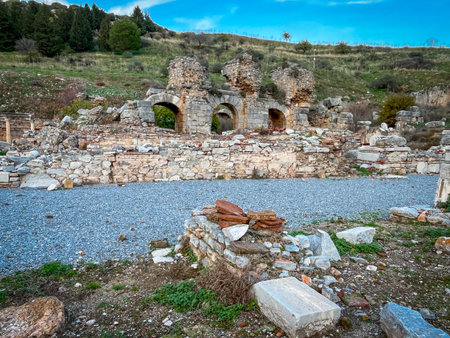 Weathered stone arches emerge from hillside ruins, framed by scattered bricks and gravel foreground. Cypress trees dot the green slope against a clear blue expanse with wispy clouds.の写真素材