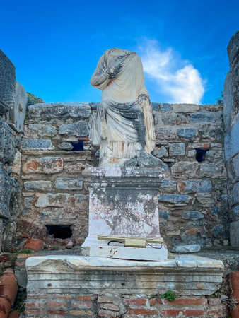 Marble statue of a female form with flowing drapery and veil over the head commands the foreground on a sturdy pedestal, framed by rugged stone walls featuring rectangular openings and terracotta fragments. Bright blue sky with scattered clouds illuminates the white marble textures, moss accents, and aged patina for a vivid classical ruin scene.の写真素材