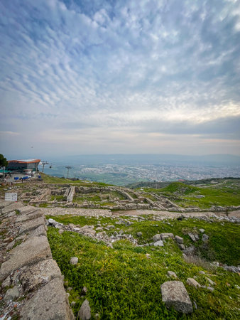 The Temple of Athena on Pergamon's Acropolis was commissioned by King Eumenes II of the Attalid Dynasty in the 2nd century BC, showcasing Hellenistic architectural grandeur. Image reveals sharp details of weathered stone ruins, vivid green overgrowth, and dramatic cloudy skies over the expansive valley.の写真素材