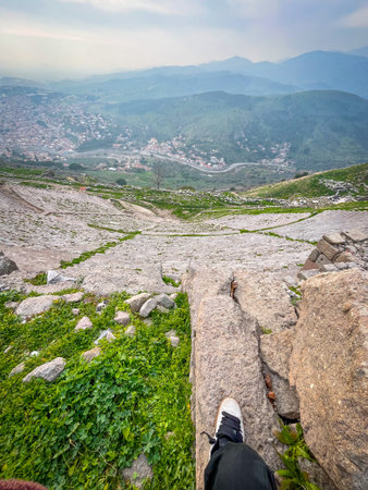 The Acropolis of Pergamon was fortified and expanded by Attalid kings like Eumenes II in the 2nd century BC, serving as the capital of the Hellenistic Kingdom of Pergamon before Roman incorporation. This high-resolution capture reveals sharp details of weathered stone terraces, vivid green ivy and grasses, against a hazy mountain skyline with crisp cityscape below.の写真素材