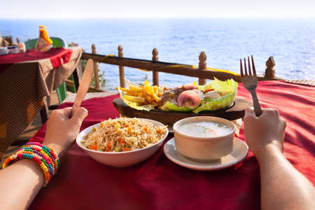 Indian vegetarian sizzler, corn soup and fried rice on the table with ocean at the background. Hands with fork and knife on the table.の写真素材