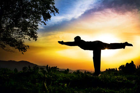 Man silhouette doing virabhadrasana III warior pose with tree nearby outdoors at sunset backgroundの写真素材