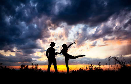 Young couple silhouette outdoors at sunset dramatic sky background. Man in cowboy hat and woman dancing near byの写真素材