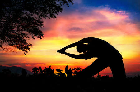 Man silhouette doing parighasana beam pose with tree nearby outdoors at sunset backgroundの写真素材