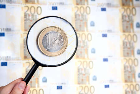 Hand holding magnifier and zooming one euro coin at two hundred euro paper currency background の写真素材