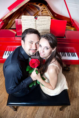 Young beautiful couple sitting on the chair near the red grand piano. Holding red rose, smiling and looking at camera の写真素材