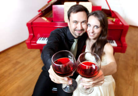 Man and woman clanging wine glasses with red liquid near by red grand pianoの写真素材